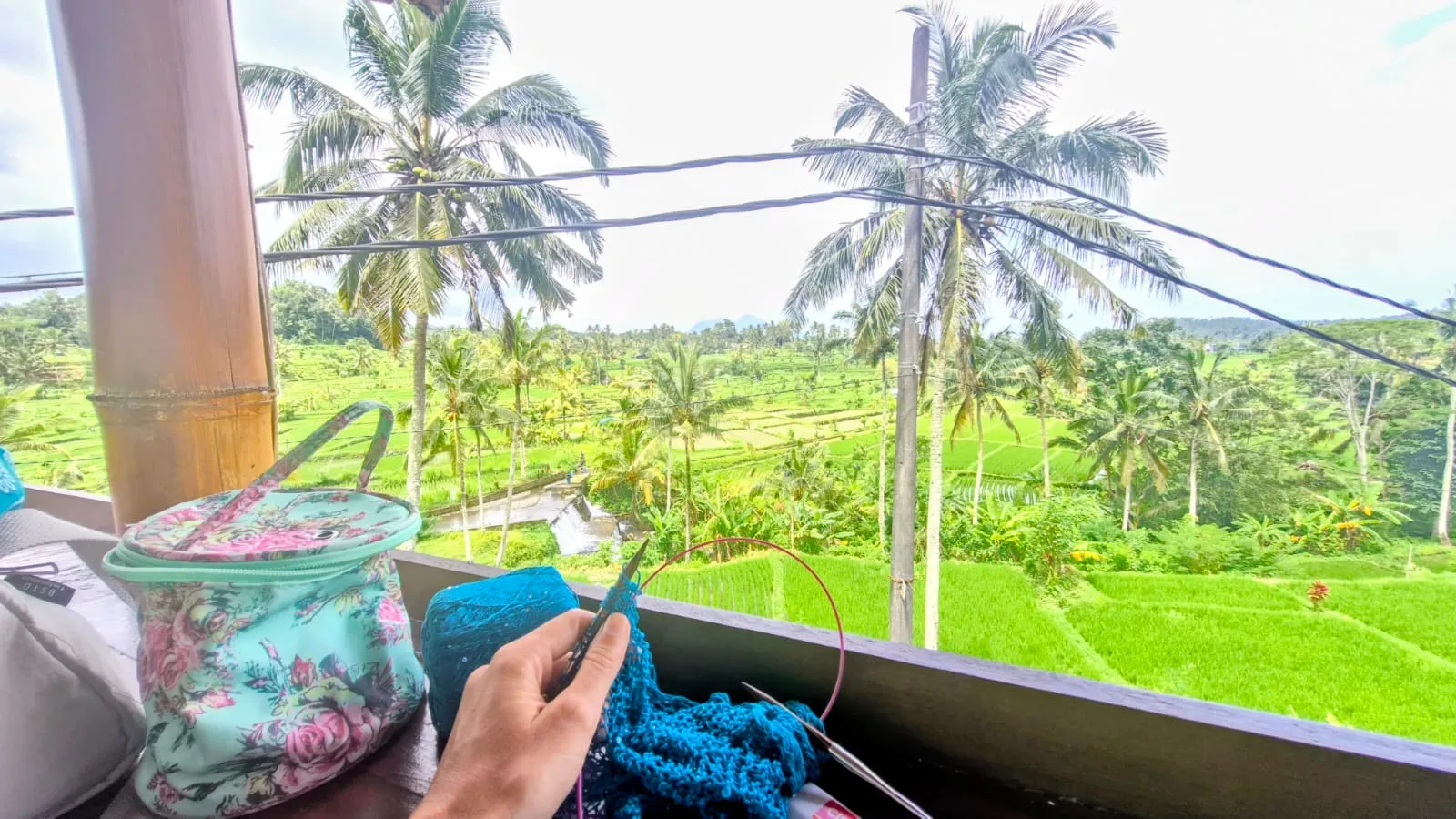 Tangan merajut benang biru di balkon, dengan sawah dan pohon kelapa di belakang.
